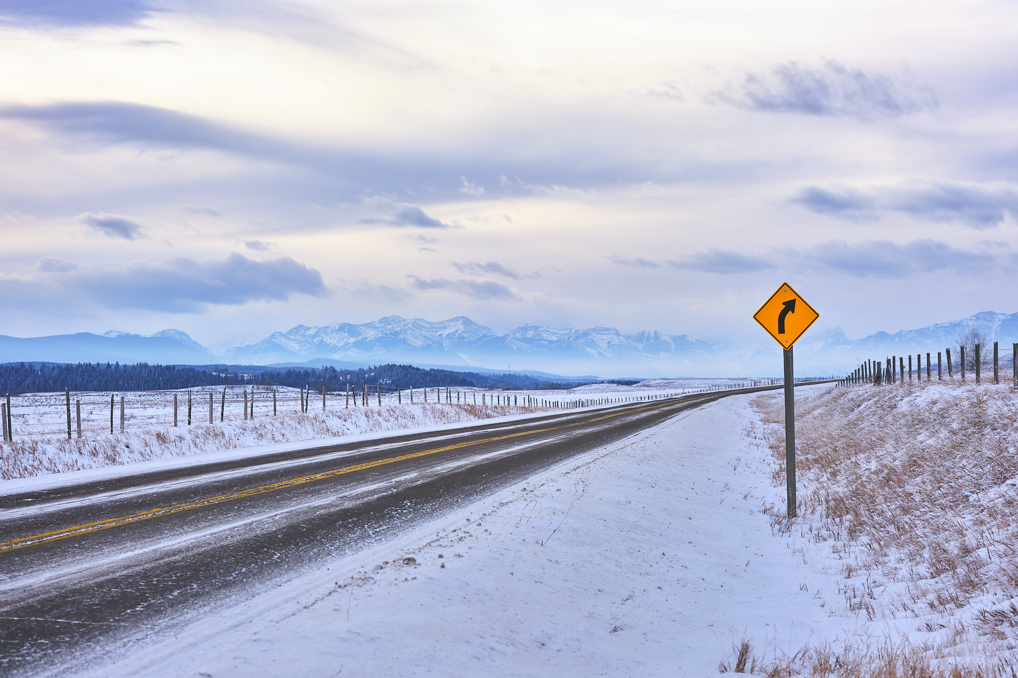 Winter highway curving toward the Rocky Mountains, fence posts and snow, southern Alberta