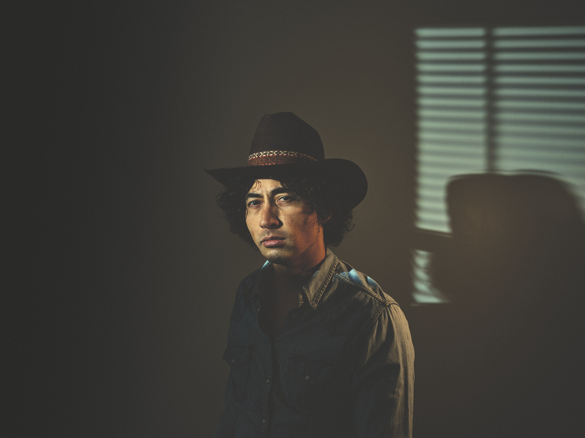 Cowboy in hat and denim, direct gaze, window blinds shadow on wall