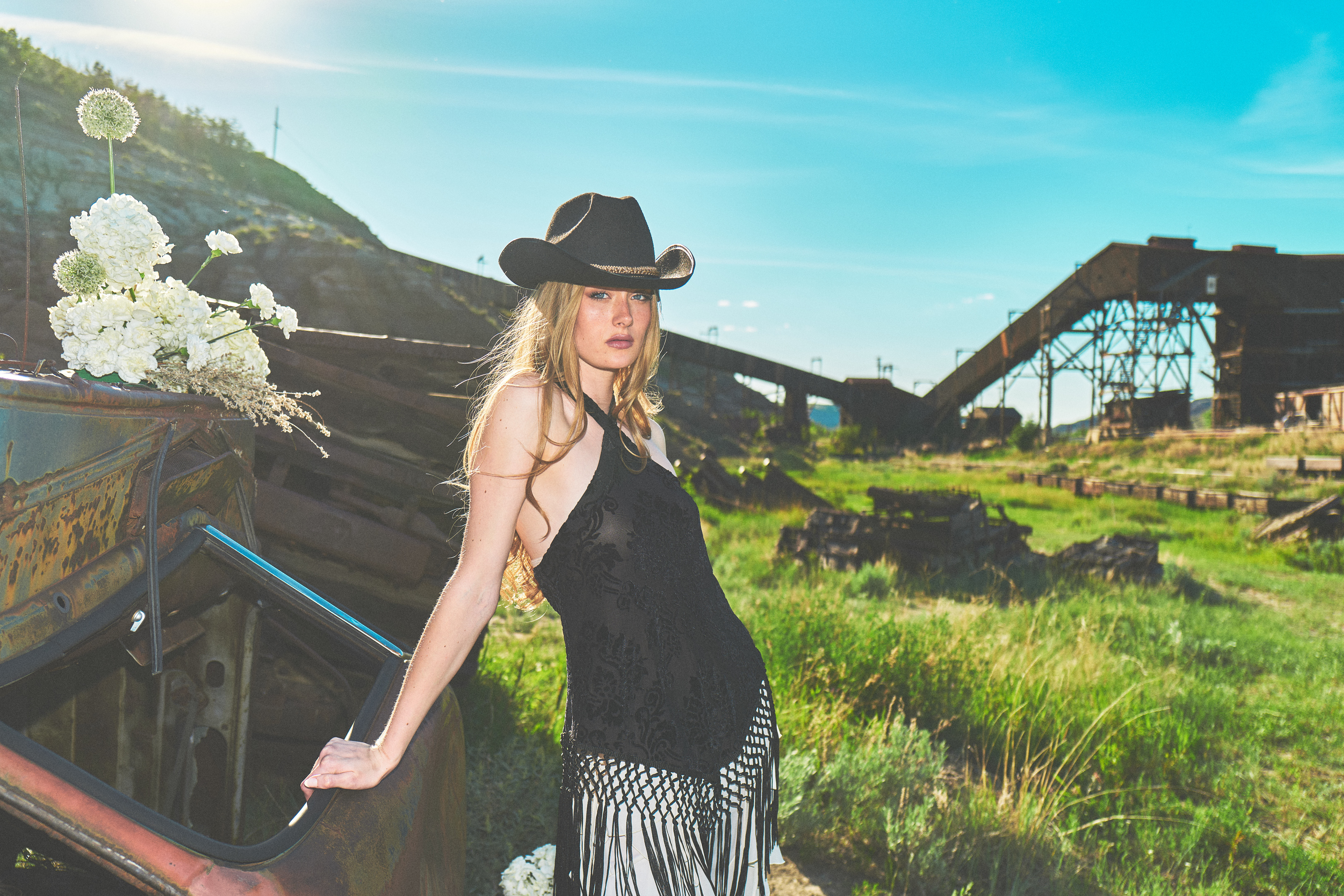 Cowgirl in black fringe dress and hat leaning on rusted truck, industrial ruins and badlands behind
