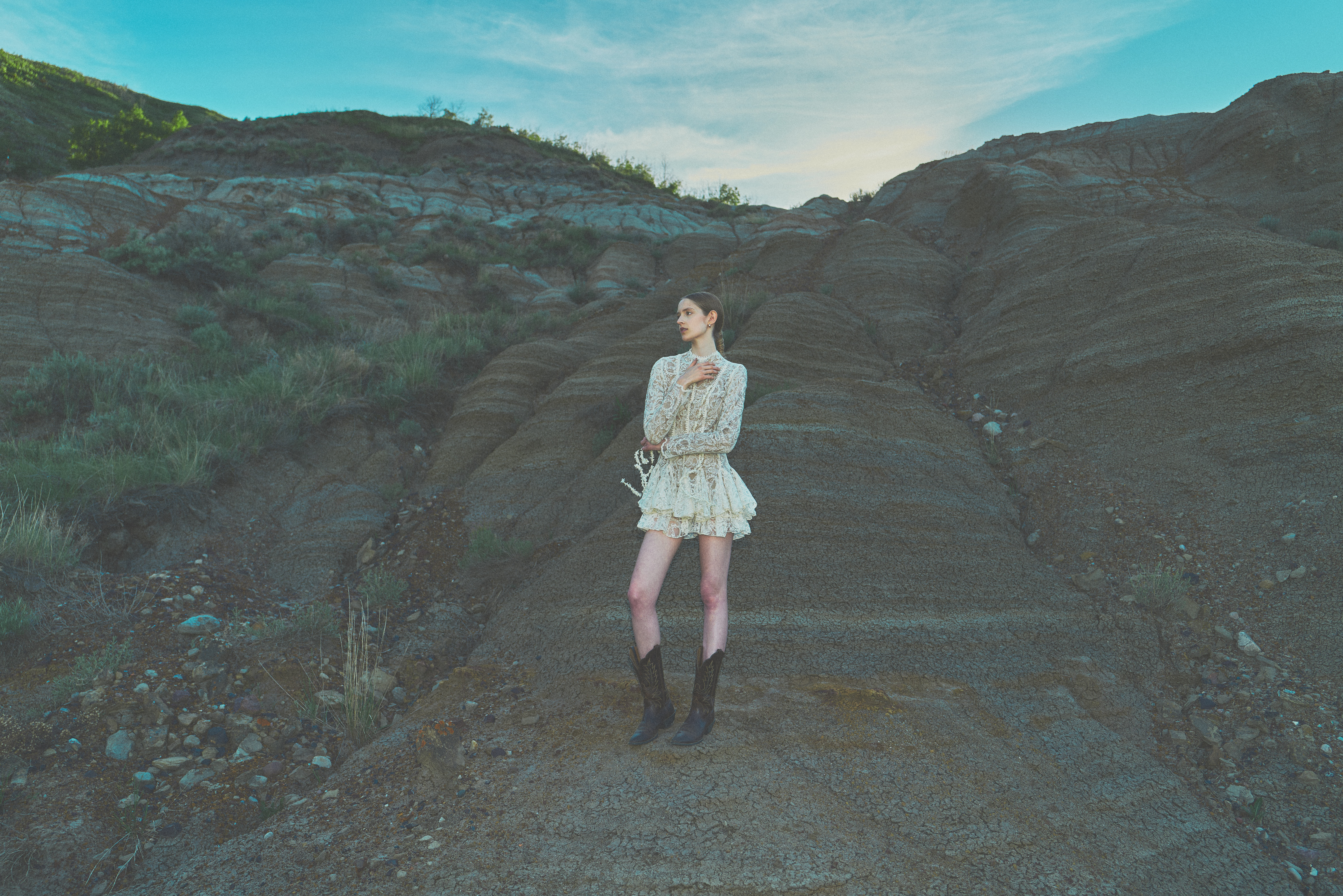 Woman in lace dress and cowboy boots standing in a badlands ravine at golden hour
