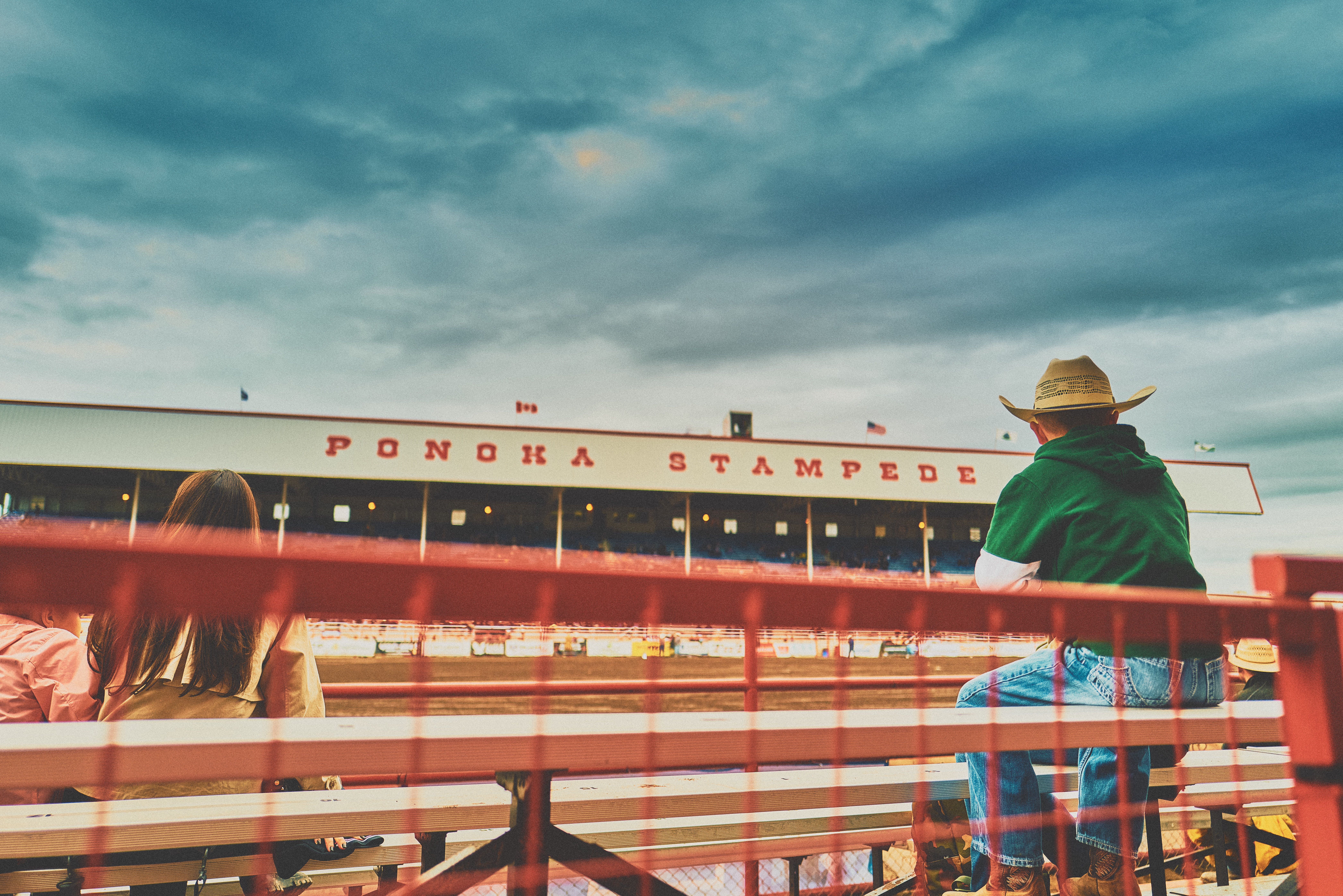 Cowboy in straw hat watching from the bleachers at Ponoka Stampede, red fence and big sky