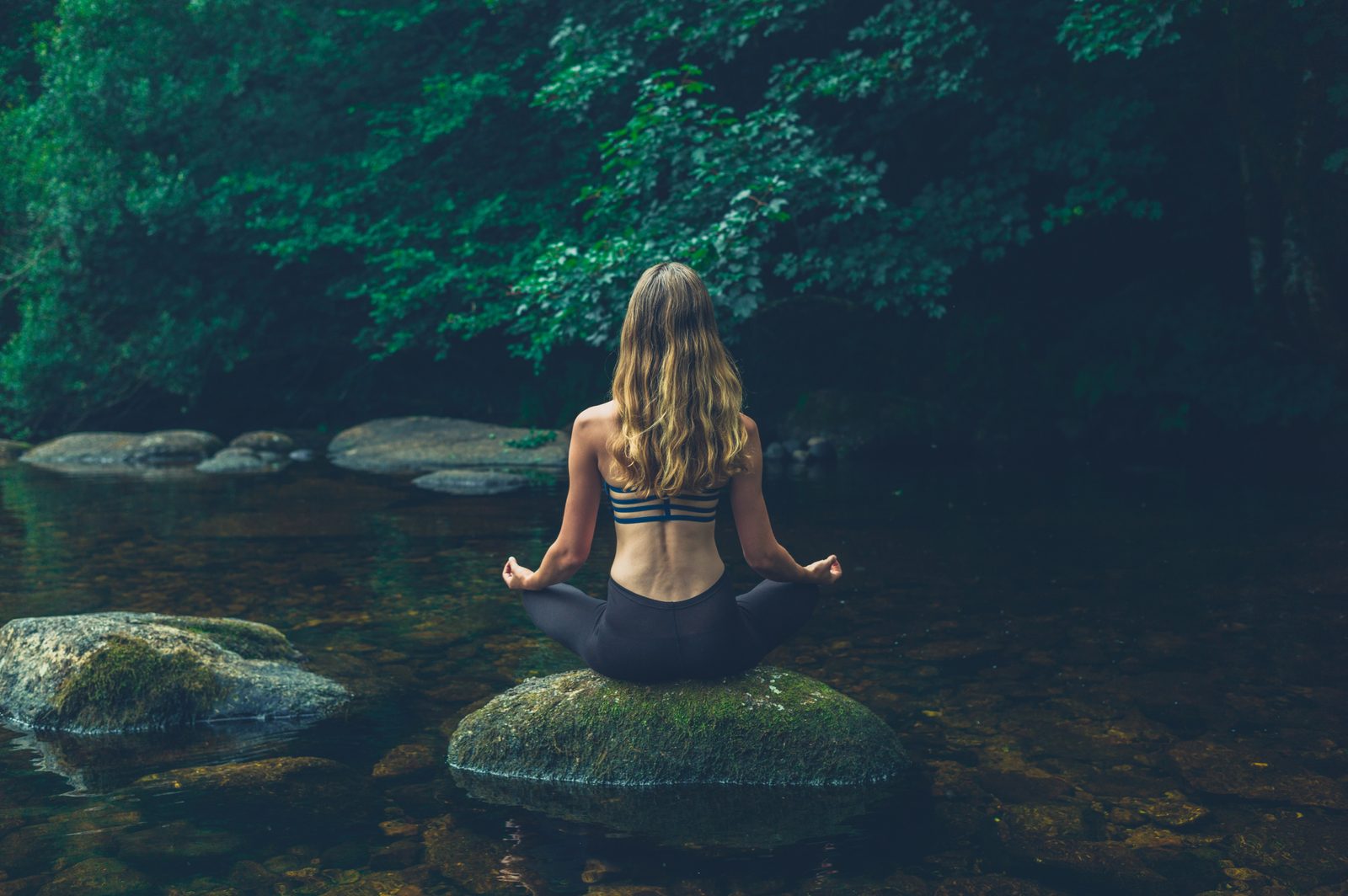 Woman meditating on rock in river — dark moody natural light photography