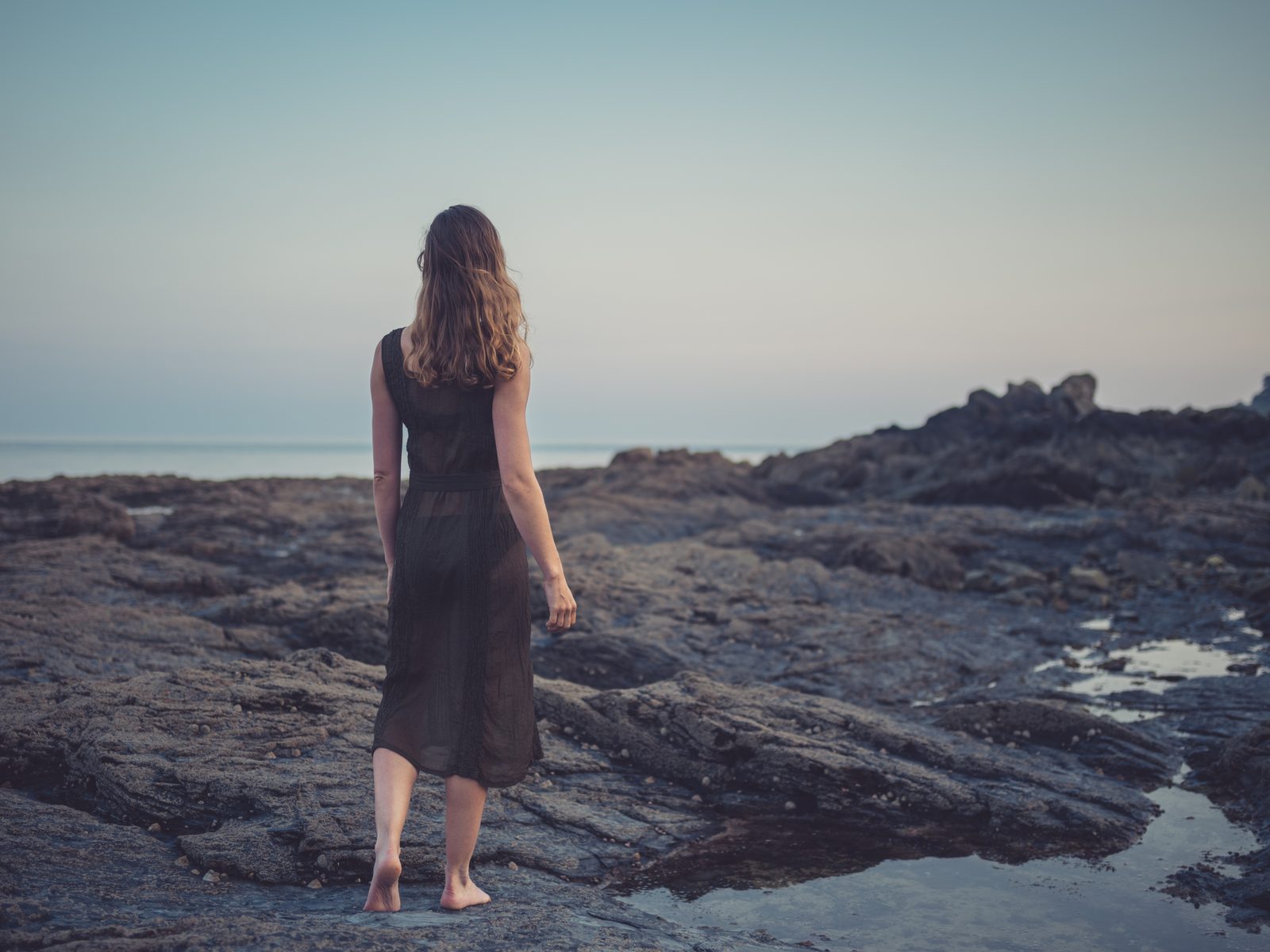 Woman walking barefoot on rocky coast at dusk — editorial portrait photography