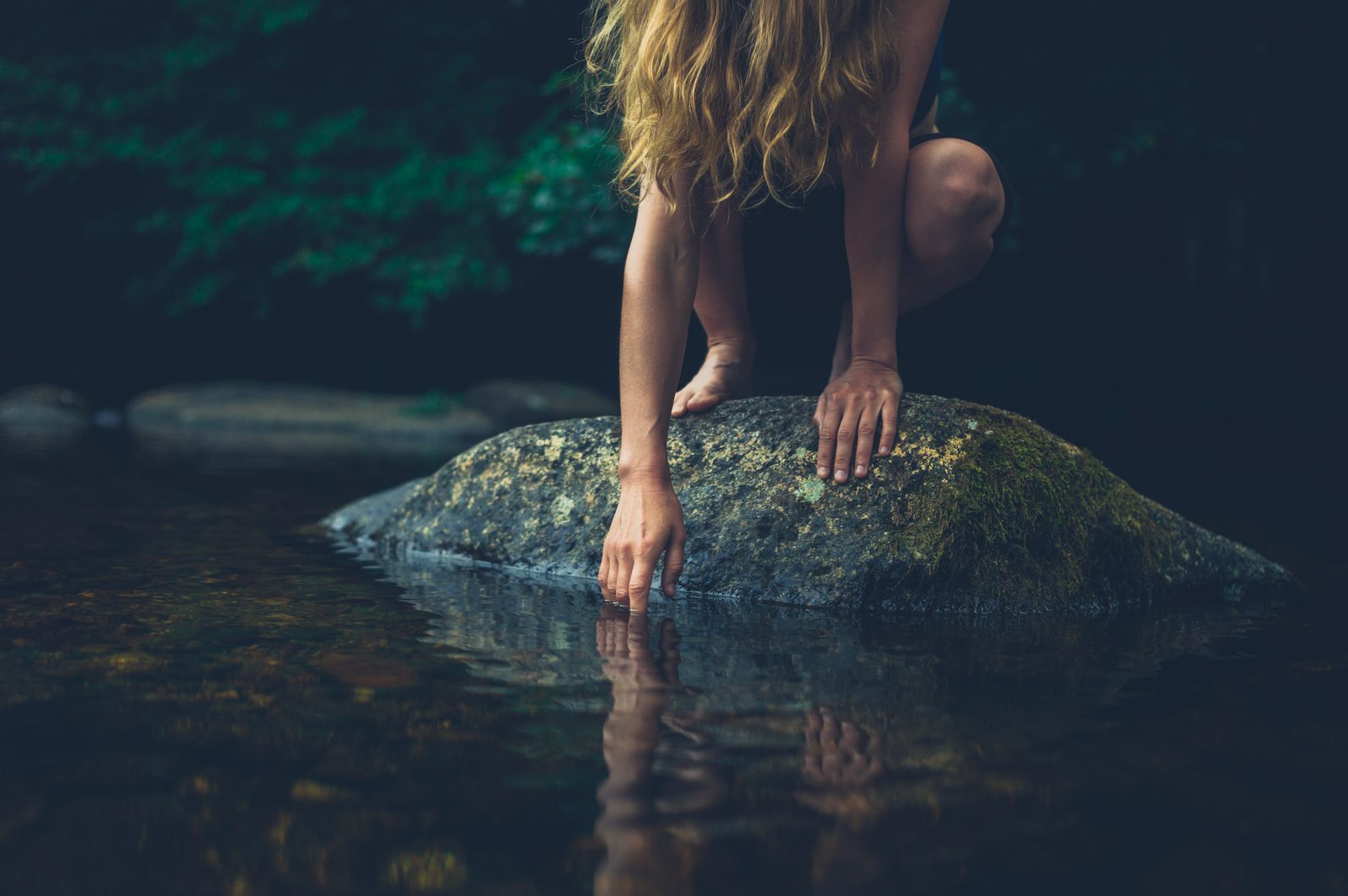 Woman sitting on mossy rock touching river water — natural light portrait