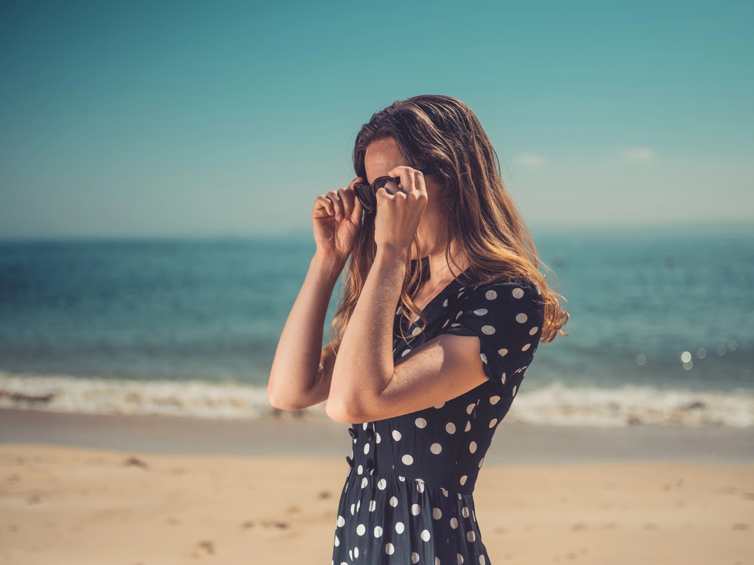 Woman in polka dot dress adjusting sunglasses on a sun-drenched beach
